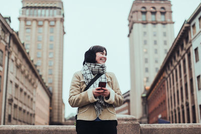 Cheerful young woman looking away while holding mobile phone against buildings in city during winter