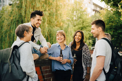 Happy young man sitting while male and female friends standing by retaining wall