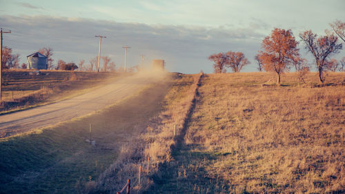 Dirt road by trees on field against sky