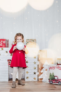 Little girl at home holding white bauble wearing red christmas dress at home over christmas deco