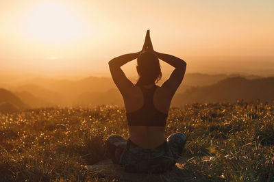 Rear view of woman meditating on field against sky during sunset