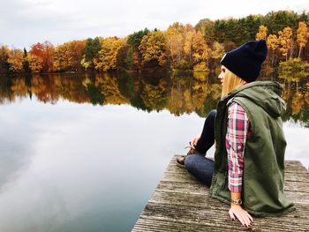 Side view of mid adult woman sitting on pier over lake against trees during autumn