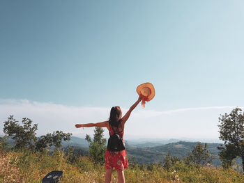Rear view of young woman with arms raised standing on mountain against blue sky during sunny day