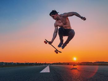 Full length of shirtless man jumping with skateboard over road against sky during sunset