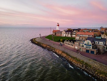 Lighthouse amidst buildings and sea against sky during sunset