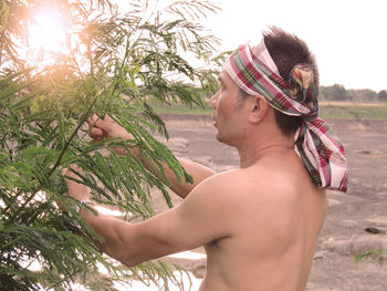 Shirtless man holding tree while standing on land against sky