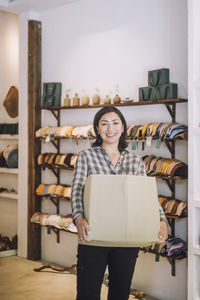 Portrait of smiling female sales clerk carrying box at fashion boutique door handle