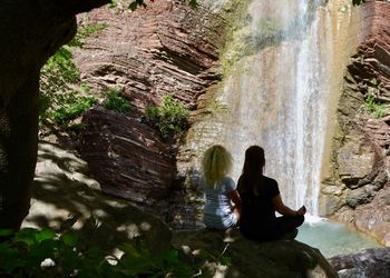 Rear view of woman sitting on rock against waterfall