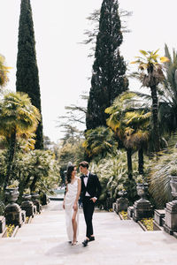 Happy lovers the bride and groom in wedding outfits walk among plants and palm trees in the old park