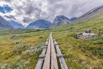 Narrow footbridge leading towards mountains against cloudy sky