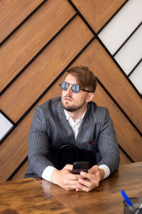 A young man in a suit and sunglasses is sitting at an office desk with a phone and looking away