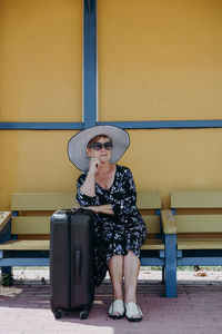 An elderly woman with a suitcase sits at a bus stop.