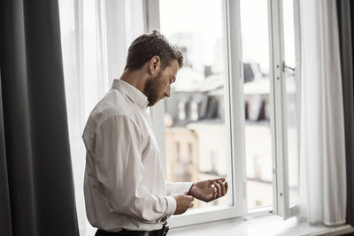 Side view of businessman getting dressed against window at hotel room