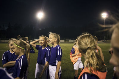 Girls standing on illuminated soccer field against sky at night