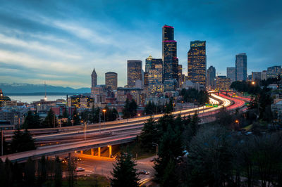 Light trails on bridge by modern buildings against sky at dusk