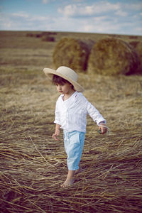 Boy a child in a straw hat and blue pants stands in a mowed field with stacks in the summer