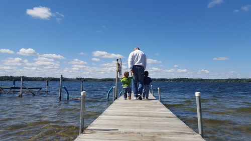 Rear view of grandfather and grandsons walking on pier amidst river against sky