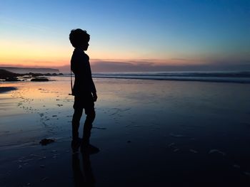 Silhouette boy standing on shore against sky during sunset