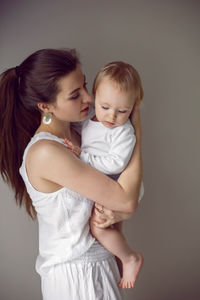 Mother holds her daughter child in her arms stands next to window in white clothes home in winter