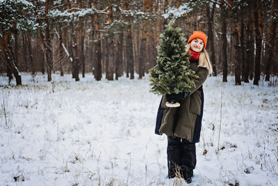 Woman in red hat holding christmas tree in snow winter park. preparing for christmas, picking