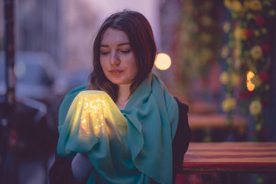 Beautiful woman holding glowing jar under blue scarf in city at dusk