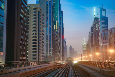 Illuminated railroad tracks amidst buildings in city against sky