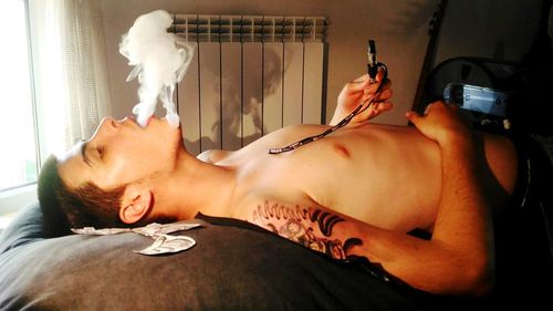 Young shirtless man smoking while lying down on bed