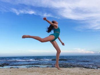 Full length of woman in bikini standing on one leg at beach against blue sky