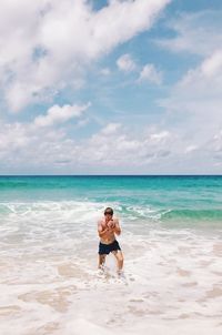 Full length of shirtless mid adult man walking at beach against cloudy sky during sunny day