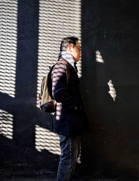 Side view of man standing against light and shadow patterned wall in city.
