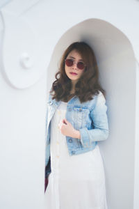 Young woman wearing sunglasses standing against white wall