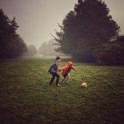 Full length of siblings playing soccer on grassy field during foggy weather