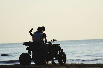 Couple with motorcycle taking selfie at beach against clear sky