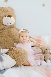 A cute one year old baby sits surrounded by soft animals toys and holds her hand on her neck