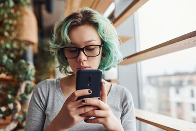 Alternative girl in glasses and with green hair sitting indoors at daytime with phone in hands.