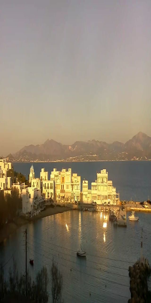 ILLUMINATED CITY BY SEA AGAINST SKY DURING SUNSET