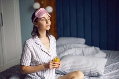 Woman in pajamas and with a sleep mask on the bed in the bedroom and drinking orange juice 