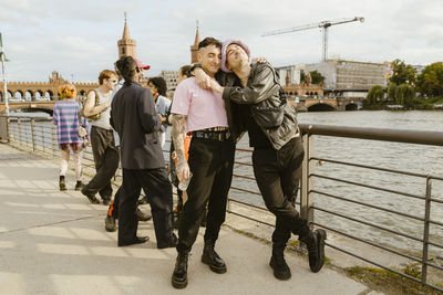 Gay man leaning on non-binary friend standing near railing on promenade in city