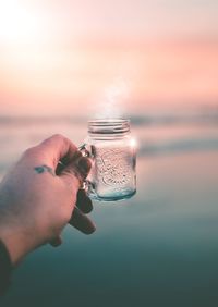 Person holding glass against sea during sunset