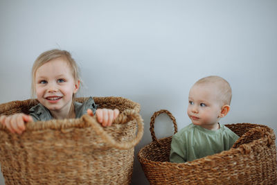 Two small children sit in wicker baskets against a white wall