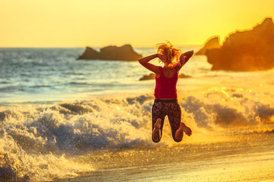 Rear view of mature woman jumping at beach against sky during sunset