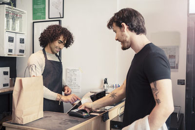 Smiling cashier using cash register while standing with male customer at checkout in organic store