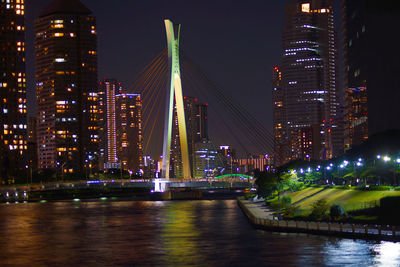 Illuminated modern buildings by river against sky at night