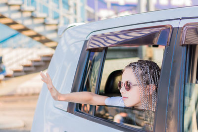 A cute teenage girl in sunglasses with afro-braids waves her hand out the window