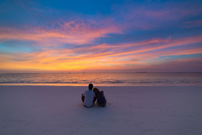 Men sitting on shore at beach against sky during sunset