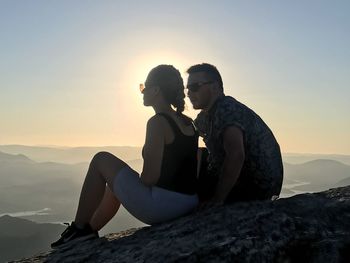 Couple sitting on rock against sky during sunset