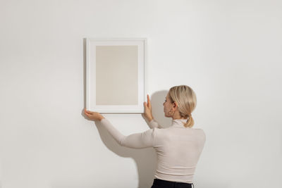 Side view of young woman hanging blank frame on the white wall.