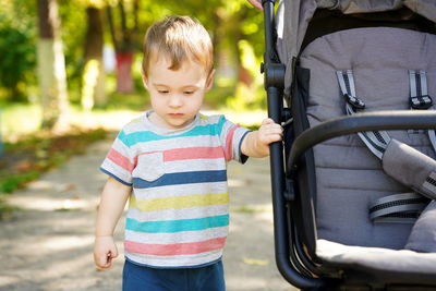 Pensive toddler boy stands near the stroller in the park. cute kid of one and a half years.