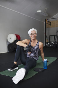 Full length portrait of happy senior woman sitting on exercise mat at health club