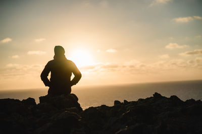 Silhouette man sitting on rock by sea against sky during sunset